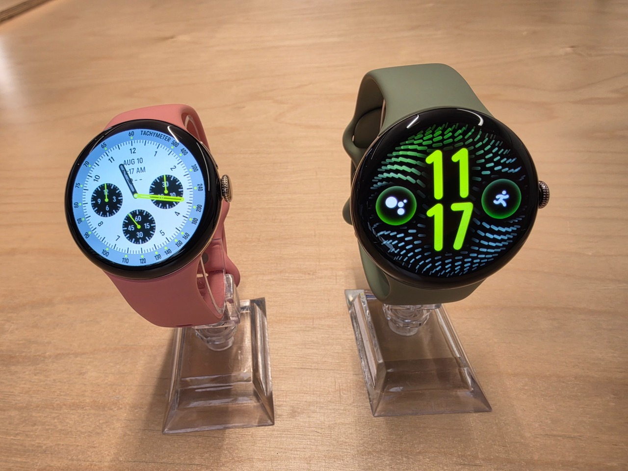 Google Pixel Watch 5 Rose cendré et Olive présentées côte à côte sur leurs supports