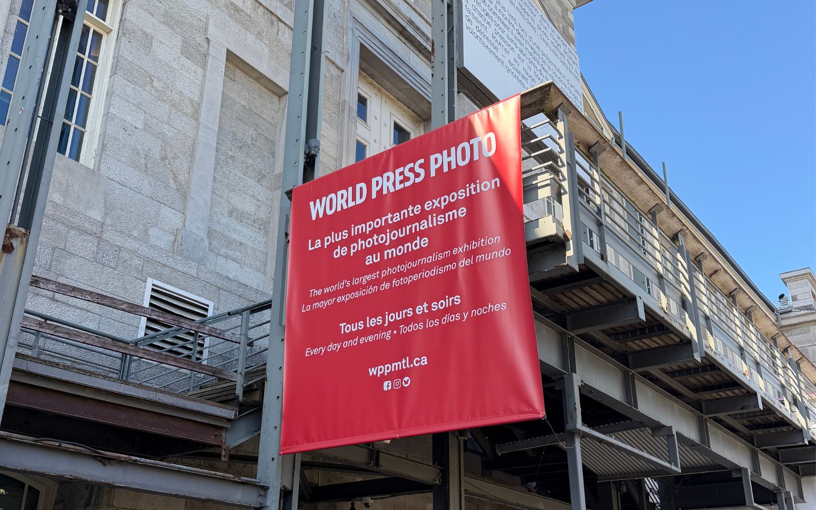 Bannière rouge de l’exposition World Press Photo accrochée à la façade d’un édifice en pierre, sous un ciel bleu